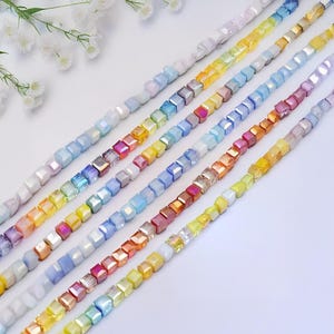 May include: Close-up of several strands of colorful, iridescent cube beads. The beads are arranged in a diagonal pattern, showcasing a spectrum of colors including pink, yellow, blue, and purple. White flowers are in the upper left corner.