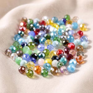 May include: A collection of faceted beads in various colors, including red, blue, green, and yellow. The beads are scattered on a light-colored, textured surface. These beads are suitable for jewelry making and craft projects.