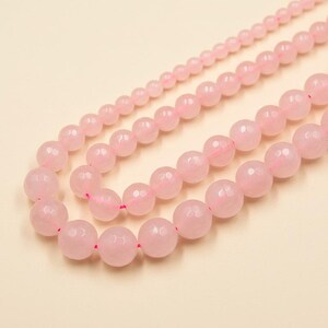 Puede incluir: Tres hilos de cuentas facetadas de cuarzo rosa claro. Las cuentas varían en tamaño, ensartadas en hilo rosa, y se muestran sobre un fondo beige claro. Las cuentas tienen una superficie pulida y reflectante.