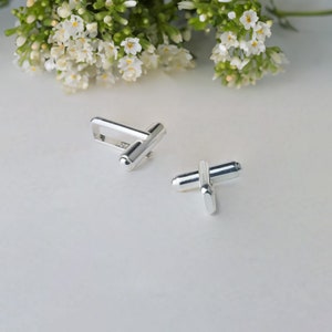 May include: A pair of silver cufflinks. One cufflink is a simple bar design, the other is a more intricate design with two bars crossing each other.