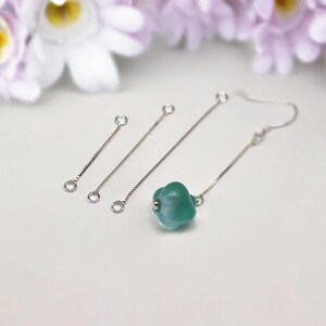 May include: A collection of silver-toned earring components, including three chain-like pieces with small loops and a single earring with a teal, flower-shaped bead. The earrings are set against a soft, blurred floral background.