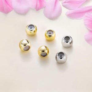 May include: Six round metal beads, three gold and three silver, with a white interior. The beads are arranged in a semi-circle on a white surface.