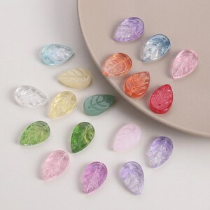 Könnte beinhalten: Eine Sammlung farbenfroher, blattförmiger Perlen in verschiedenen Farbtönen, darunter transparent, blau, gelb, grün, rosa, orange, rot und lila. Jede Perle hat ein detailliertes Blattader-Design. Die Perlen sind auf einem neutralfarbenen Teller angeordnet.