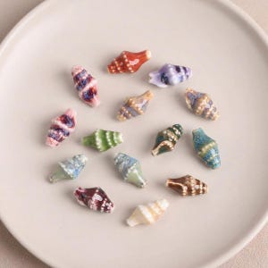 May include: A selection of small, ceramic seashells in a variety of colours, including red, purple, green, and blue. The shells are displayed on a white plate, highlighting their intricate designs and glossy finish.
