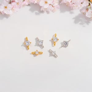 Pode incluir: Uma variedade de pequenas e brilhantes peças de joalharia, incluindo pingentes e um anel, em tons de ouro e prata. Cada peça apresenta uma gema clara e redonda. Os artigos estão dispostos sobre uma superfície branca, com delicadas flores cor-de-rosa ao fundo.