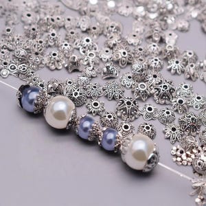 May include: A strand of beads with silver-toned metal accents and white and blue pearl-like beads. The beads are scattered around the strand, showing a variety of designs.