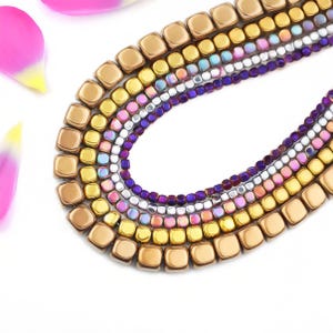 May include: A close-up of a multi-strand necklace with square and round beads in shades of gold, purple, pink, and iridescent colors. The necklace is arranged in a curved shape against a white background. Pink and yellow flower petals are in the upper left corner.