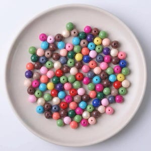 Op de afbeelding: Een verzameling kleurrijke ronde kralen in verschillende tinten groen, roze, blauw, bruin en geel, gerangschikt op een lichtbeige bord. De kralen zijn uniform van grootte en vorm, perfect voor het maken van sieraden of knutselen.