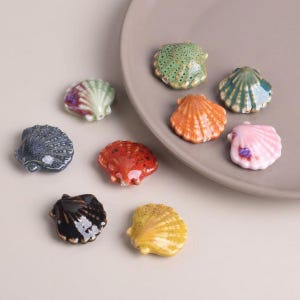 May include: A collection of ceramic seashell figurines in various colors, including green, orange, pink, and blue. The seashells are arranged on a neutral-toned plate and surface, showcasing their detailed textures and glossy finishes. These decorative pieces add a coastal theme.