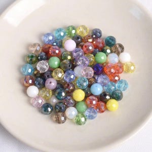 Könnte beinhalten: Eine Sammlung farbenfroher, facettierter Perlen in verschiedenen Größen und Farbtönen, darunter Blau, Grün, Gelb, Rosa und Klar. Die Perlen sind auf einem weißen, flachen Teller verstreut, ideal für Schmuckherstellung oder Basteln.