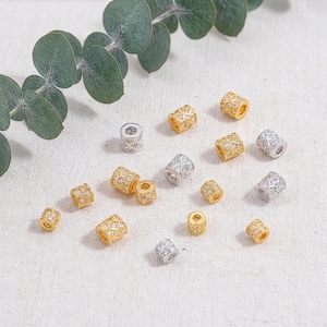 Puede incluir: Un surtido de cuentas decorativas en tonos dorados y plateados. Las cuentas cilíndricas presentan un diseño estampado y pequeños detalles brillantes. Algunas cuentas están dispersas sobre una superficie blanca, con follaje verde en el fondo.