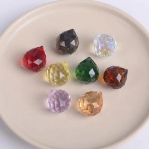 May include: A selection of multi-coloured faceted crystal pendants, including red, brown, green, and yellow. The crystals are displayed on a light beige plate, highlighting their geometric shapes and reflective surfaces. These decorative crystals are suitable for jewellery making or home decor.