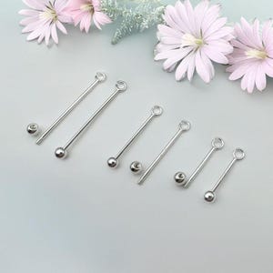 Peut inclure: Six bijoux industriels en argent, de type barbell, avec une boule à une extrémité et un petit anneau à l'autre. Les bijoux sont présentés sur une surface bleu clair avec des fleurs roses en arrière-plan.