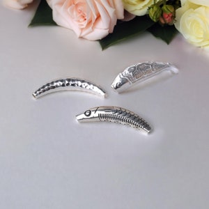 May include: Three silver fish-shaped decorative objects. One has a hammered texture, while the others feature detailed engraved designs. The objects are arranged on a white surface, with soft pink and yellow roses in the background.