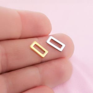 Op de afbeelding: Twee kleine, rechthoekige sieradenonderdelen. Eén is goudkleurig, de andere zilverkleurig. Beide hebben een hol, open midden. De onderdelen worden in de hand van een persoon gehouden tegen een zachtroze achtergrond.