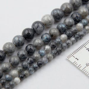 May include: Close-up of strands of grey and blue gemstone beads. The beads have a faceted surface and are arranged in rows. A ruler is placed next to the beads for size comparison. The beads are likely for jewellery making.