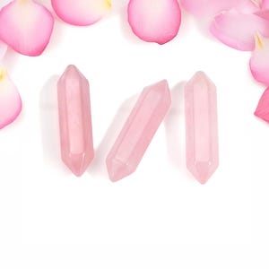 May include: Three pink crystal points, also known as rose quartz, are arranged on a white background. The crystals are smooth and polished, with a pointed tip and a flat base.