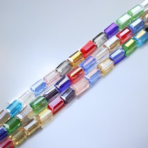May include: A collection of colourful, rectangular glass beads. The beads are arranged in a diagonal line, with the colours ranging from clear to red, blue, green, and gold.
