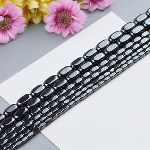 Könnte beinhalten: Eine Reihe schwarzer Hämatitperlen, jede Perle ist oval geformt und hat eine glatte, polierte Oberfläche. Die Perlen sind in Reihen angeordnet und bilden ein optisch ansprechendes Muster.