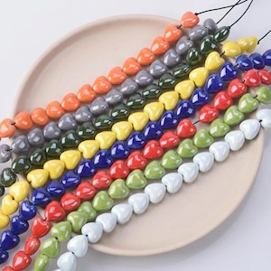 May include: Strands of heart-shaped beads in various colors, including orange, gray, green, yellow, blue, red, green, and white. The beads are strung on black cords and arranged on a beige plate. These beads are suitable for jewelry making.