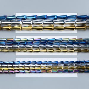 May include: Close-up of rows of metallic cone-shaped beads in blue, silver, and gold, arranged on white cards. The beads have a faceted surface, creating a shimmering effect. These beads are likely for jewellery making, with a variety of colours.