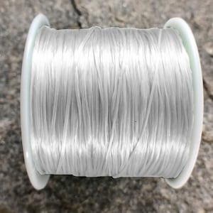 Op de afbeelding: Een spoel met wit elastisch koord, strak gewikkeld op een witte plastic spoel. Het koord is glad en glanzend en lijkt sterk en duurzaam.