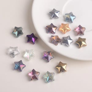 Puede incluir: Una colección de cuentas en forma de estrella en varios colores, incluyendo transparente, iridiscente, dorado, plateado y morado. Las cuentas están dispersas sobre una superficie blanca y cerca de un plato blanco. Estas cuentas son adecuadas para la fabricación de joyas y manualidades.