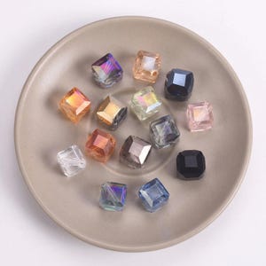May include: A collection of faceted, square beads in various colors, including orange, purple, clear, and black, are arranged on a neutral-toned plate. The beads have a reflective, iridescent quality, suggesting they are made of glass or crystal.