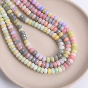 May include: Strands of colorful gemstone beads in pastel shades of pink, yellow, green, gray, and purple. The beads are arranged on a beige plate, showcasing their smooth, rounded shapes. These beads are ideal for jewelry making.