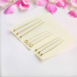 Peut inclure: Un ensemble de dix épingles à tête dorées avec différents styles de tête. Les épingles à tête sont disposées en deux rangées sur une surface blanche.