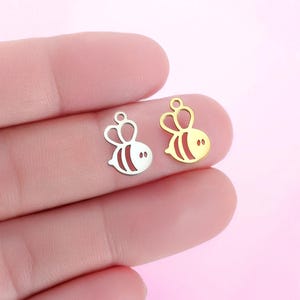 May include: Two small bee-shaped charms, one silver-colored and one gold-colored, are displayed. Each charm features a crown, a rounded body with striped details, and small eye details. The charms are held between fingers, showcasing their size and design.