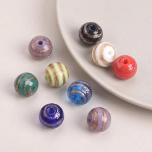 Puede incluir: Una colección de nueve cuentas de vidrio esféricas en varios colores, incluyendo negro, blanco, rojo, verde, azul y morado. Cada cuenta tiene un orificio central y está decorada con una banda dorada brillante en espiral por la superficie.