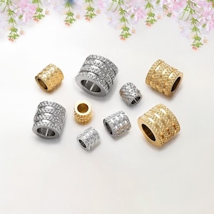 Puede incluir: Una colección de cuentas de metal dorado y plateado con un diseño texturizado. Las cuentas son de varios tamaños y están dispuestas sobre una superficie blanca.