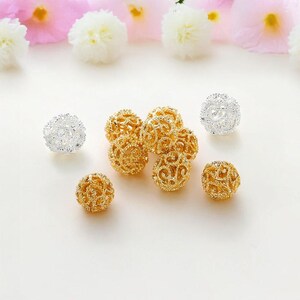May include: Close-up of several filigree orb beads in gold and silver. The beads have an open, lacy design. The background is a soft white with pink and white flowers.