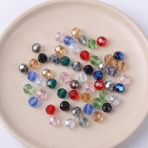 May include: A collection of faceted beads in various colors, including red, green, blue, and clear. The beads are round and have a shiny, reflective surface. They are scattered on a light beige plate, suitable for jewelry making or crafts.