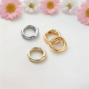 Pode incluir: Três chaveiros de prata e ouro com fecho giratório. Os chaveiros estão sobre uma superfície branca.