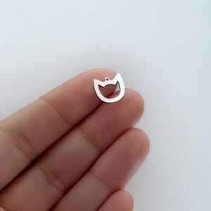 May include: A small, silver-colored cat-shaped charm with a cutout face and a small hole at the top for stringing. The charm is smooth and shiny, perfect for jewelry making or crafting projects.