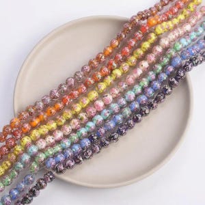 May include: Strands of colorful glass beads with floral patterns. The beads are arranged in a rainbow spectrum, showcasing a variety of colors including red, orange, yellow, pink, blue, and purple. Each bead is approximately 0.4 inches in diameter, perfect for jewelry making.