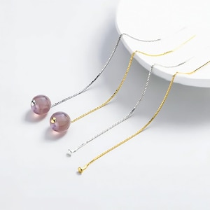 Puede incluir: Elegantes pendientes colgantes con un orbe púrpura translúcido y una delicada cadena. Disponibles en tonos plateados y dorados. La cadena mide aproximadamente 7.6 cm de largo, con una pequeña estrella o bola al final.