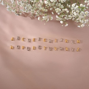 Puede incluir: Una colección de colgantes de alfabeto dorados y plateados, dispuestos sobre una superficie rosa. Los colgantes, en una fuente sans-serif, están dispersos por la superficie. Flores blancas en la parte superior de la imagen.