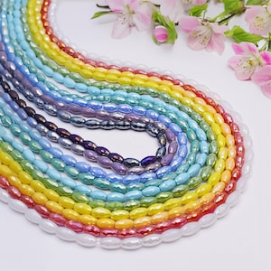 Puede incluir: Una colección de cuentas ovaladas y coloridas dispuestas en un patrón de arcoíris. Las cuentas varían en color desde blanco hasta rojo, naranja, amarillo, verde, azul y morado. Flores rosas en el fondo.