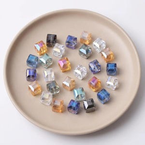 Op de afbeelding: Een verzameling facetgeslepen, kubusvormige kralen in diverse kleuren, waaronder helder, amber, blauw en iriserend. De kralen zijn gerangschikt op een beige bord, waardoor hun reflecterende oppervlakken worden benadrukt. Ideaal voor het maken van sieraden of knutselprojecten.