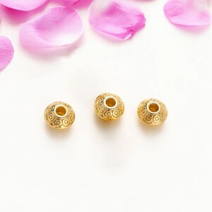 May include: Three gold-colored beads with intricate spiral patterns and a central hole. The beads are arranged on a white surface, with pink rose petals scattered above.