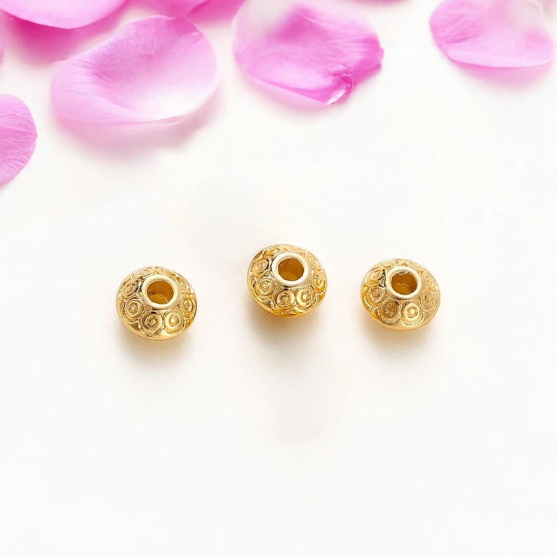 May include: Three gold-colored beads with intricate spiral patterns and a central hole. The beads are arranged on a white surface, with pink rose petals scattered above.