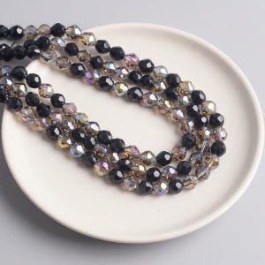 Puede incluir: Tres hilos de cuentas de cristal negro e iridiscente facetadas están dispuestas sobre un plato blanco. Las cuentas tienen una cualidad reflectante, captando la luz para mostrar una gama de colores. Las cuentas son probablemente para la fabricación de joyas.