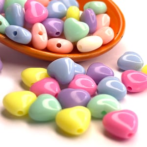 May include: A collection of pastel-colored heart-shaped beads. The beads are in shades of pink, blue, yellow, and green. The beads are scattered on a white surface.