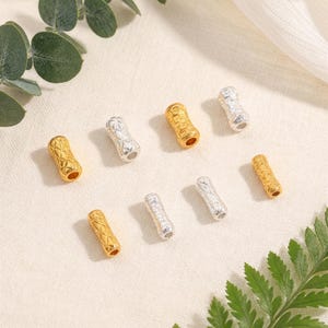 May include: An assortment of gold and silver-colored metal beads. The beads have a textured, quilted pattern and a cylindrical shape. The beads are scattered on a light-colored surface, with green foliage in the background. These beads are suitable for jewelry making.