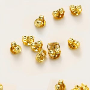 May include: A close-up of a group of gold earring backs. The backs are designed to help keep earrings in place.