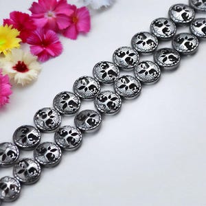 May include: Rows of silver-toned, round buttons with a tree-of-life design. The buttons have a black, recessed pattern and a shiny, metallic finish. The buttons are arranged in parallel lines, suggesting they are for sewing or crafting.