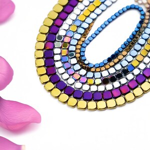 以下が含まれることがあります： 金色、紫色、青色、銀色など、さまざまな虹色の色をした正方形のヘマタイトビーズの糸。ビーズは、一番下のビーズが大きく、一番上のビーズが小さい、段階的なパターンで配置されています。